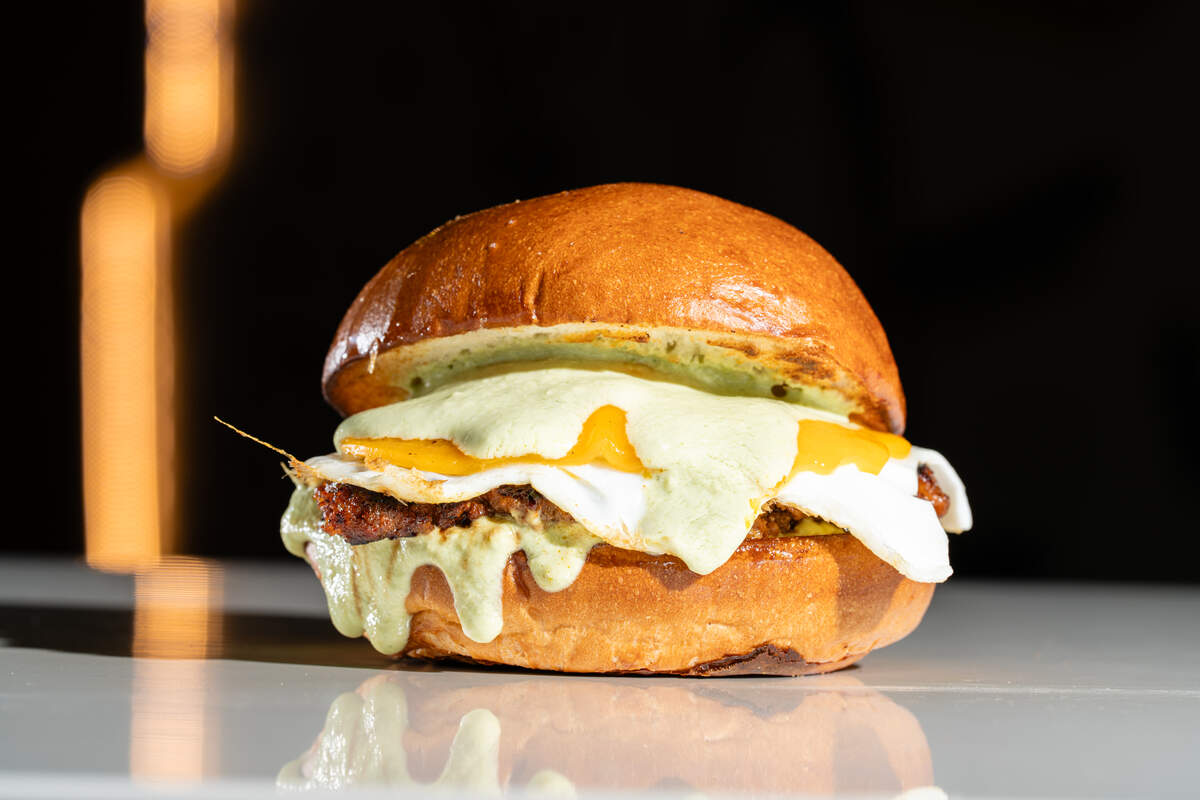 An Eggbred breakfast sandwich features the house pork chorizo patty, served with an over medium egg, sharp cheddar, avocado-cilantro crema on a toasted milk bun.